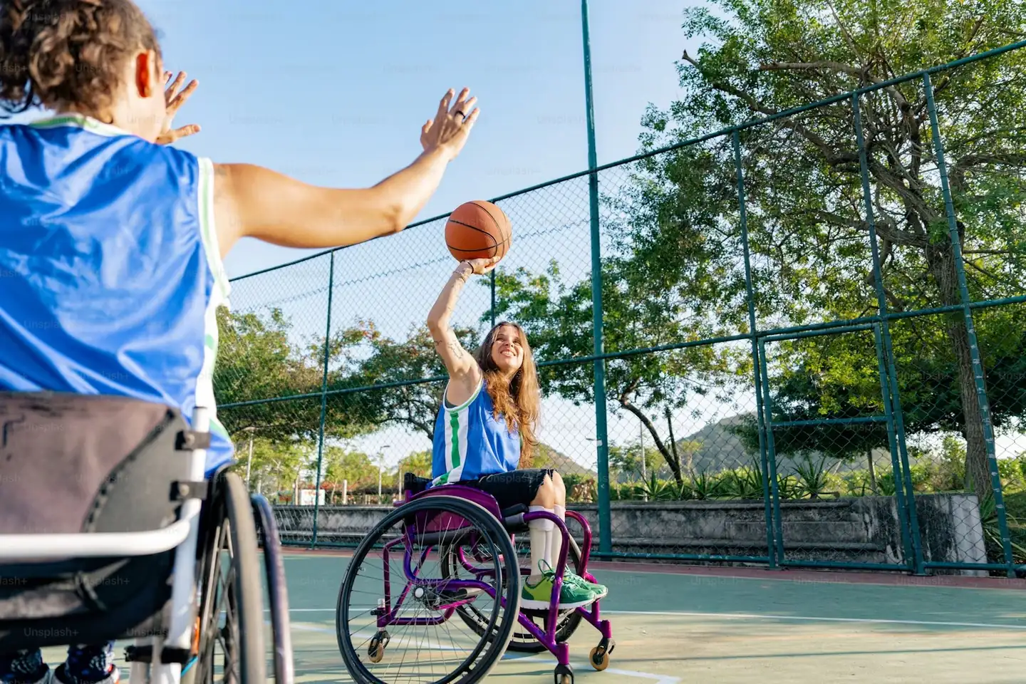Two women in wheelchairs playing basketball on a community court, one shooting for the hoop